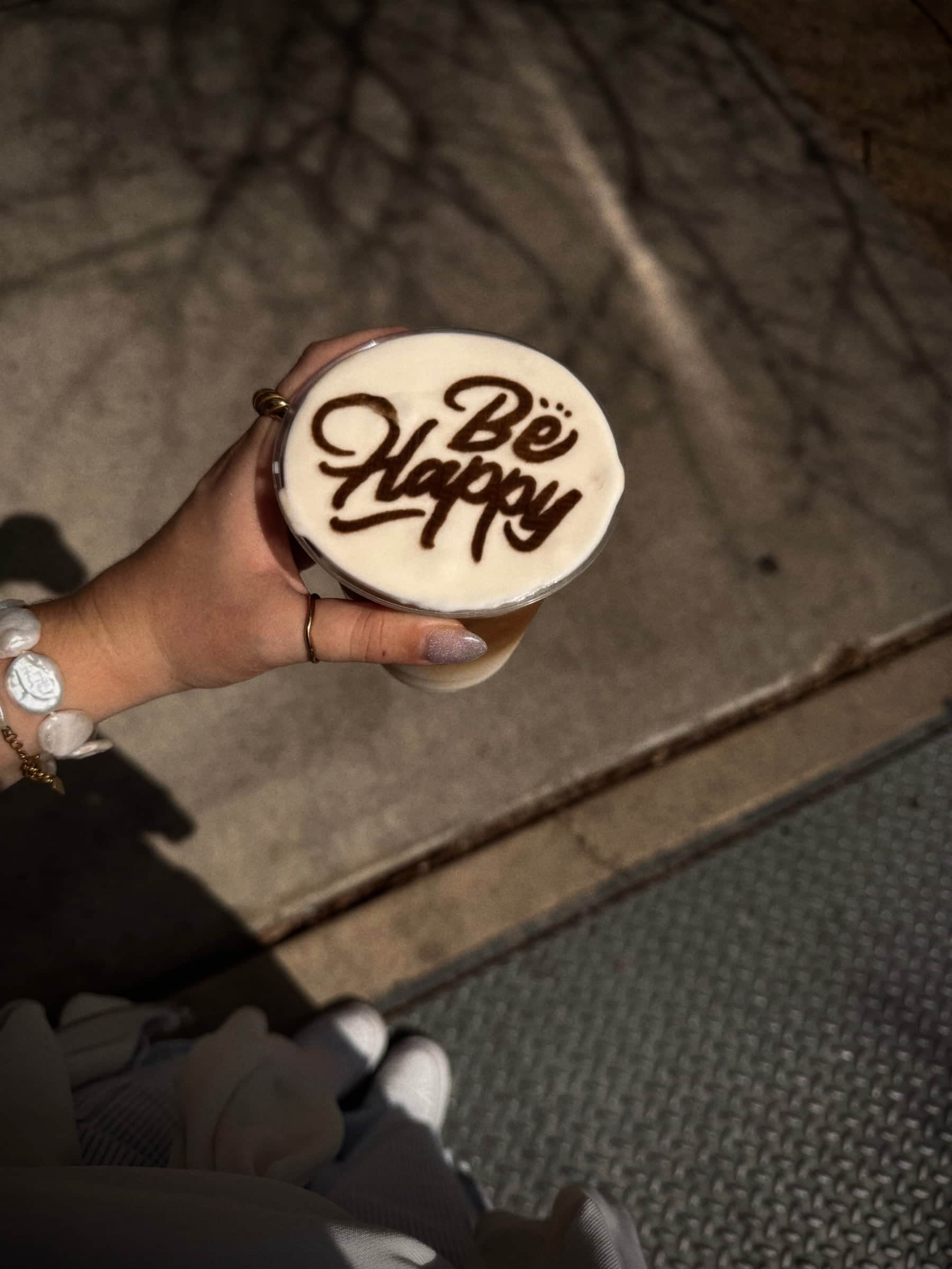 Hand holding a latte with Be Happy latte art against a dark wood floor