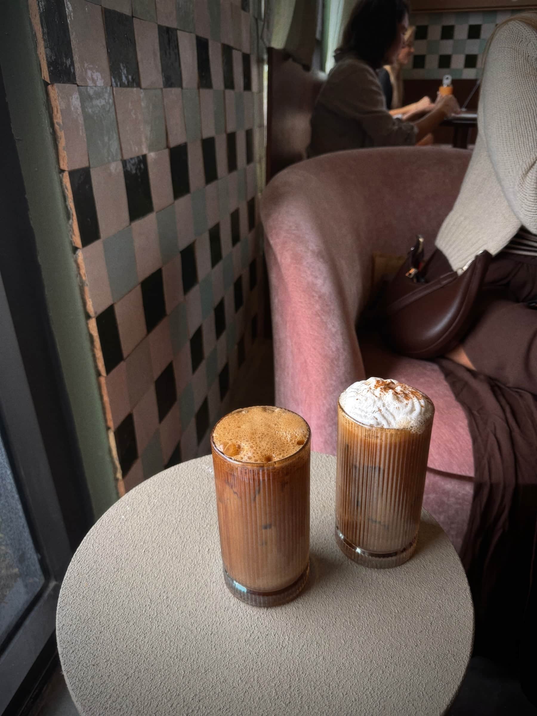Two iced mochas topped with whipped cream on a round table beside a pink velvet chair