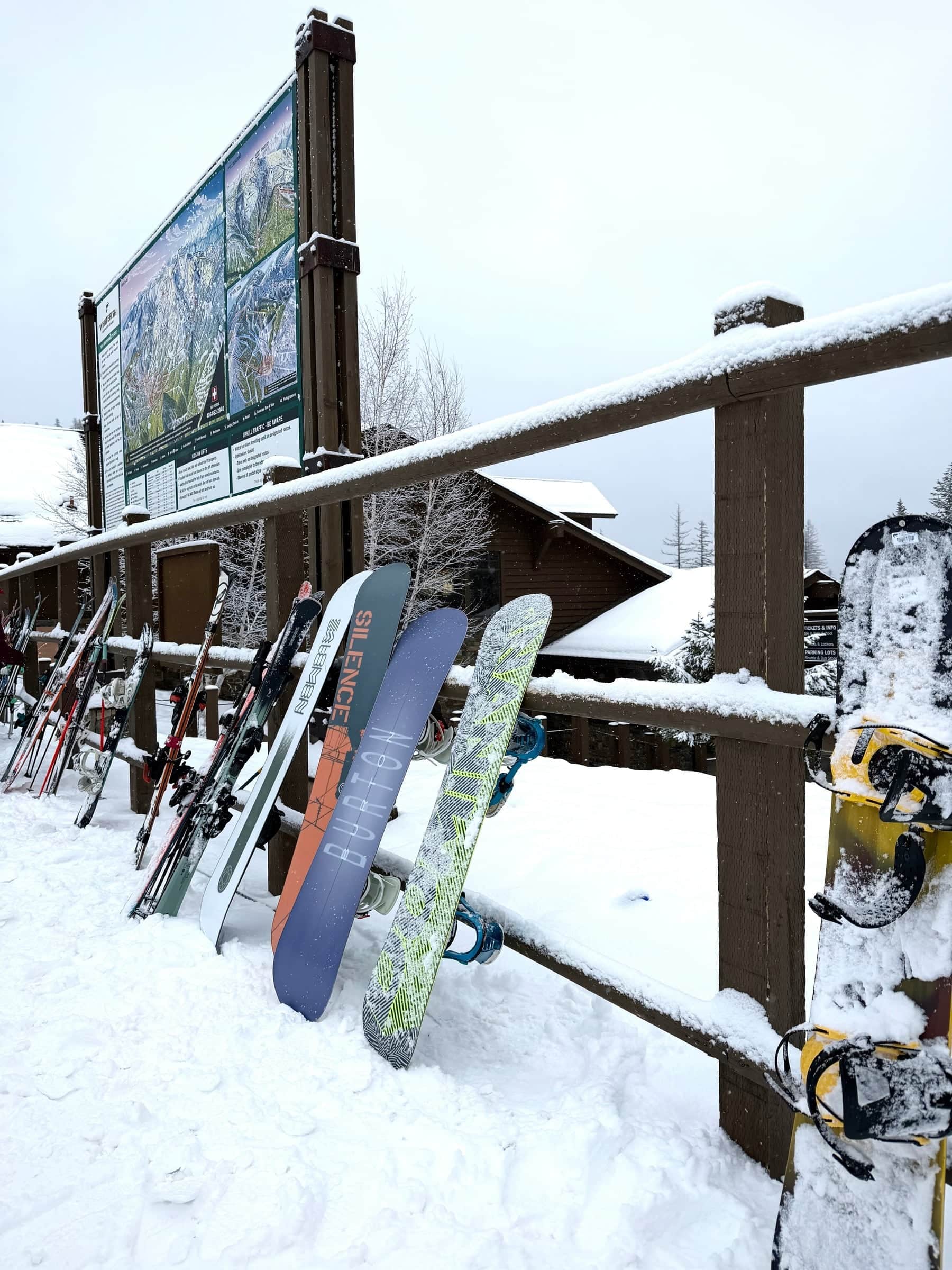 Skis and snowboards lined up on a rustic wooden rack in fresh snow