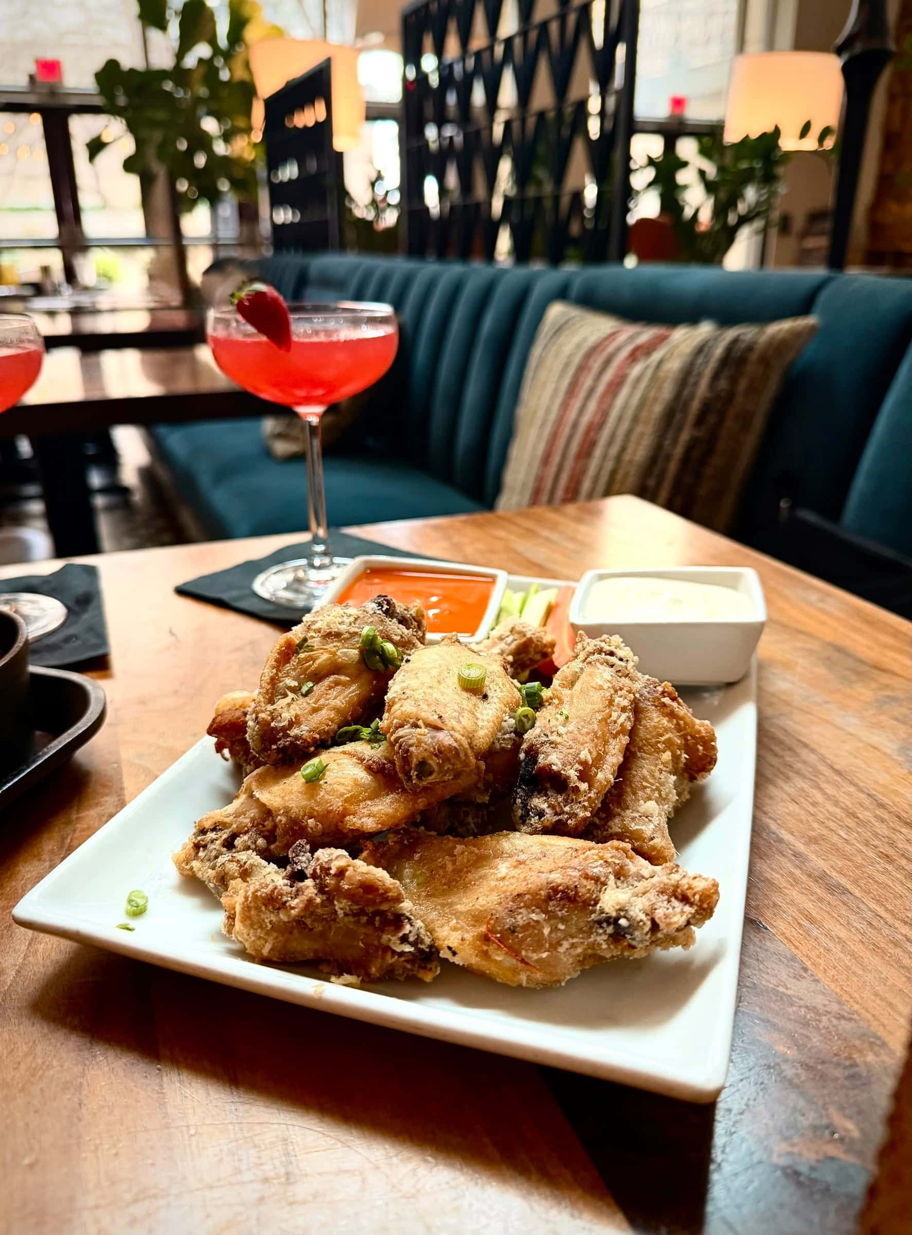 Crispy wings with dipping sauces and a pink cocktail on a table beside a teal velvet booth at Ambler