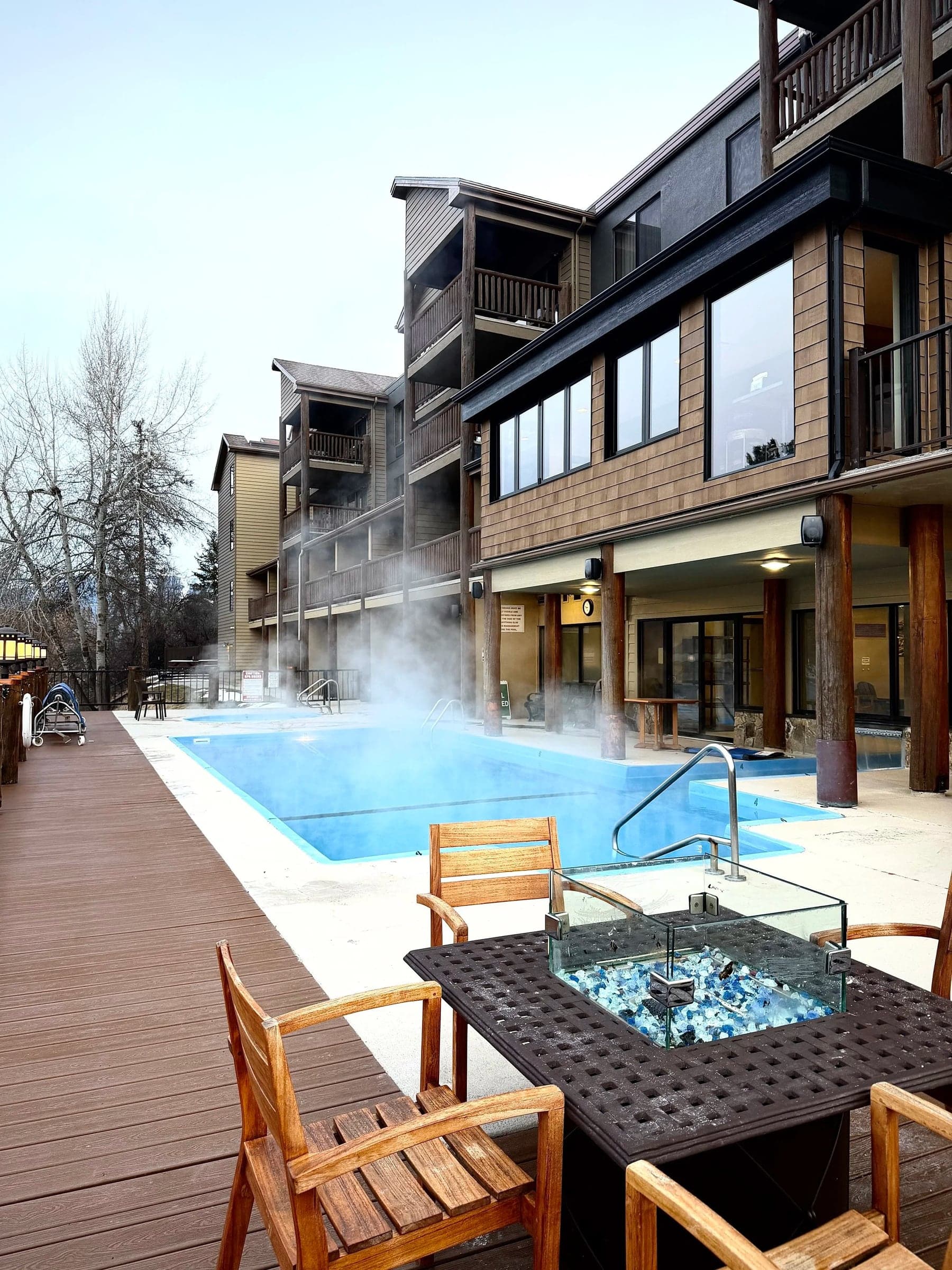 Steam rising from the heated outdoor pool at Whitefish Riverfront Resort, with timber balconies and a fire table on the deck